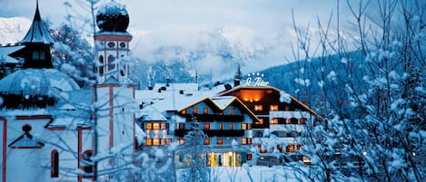 Un hotel cubierto de nieve con varias construcciones, incluyendo una torre destacada y un amplio edificio principal con ventanas iluminadas.