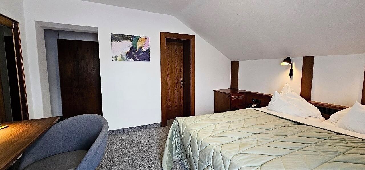 A hotel room with a bed, a desk, a chair, a nightstand, and a wall-mounted lamp.
