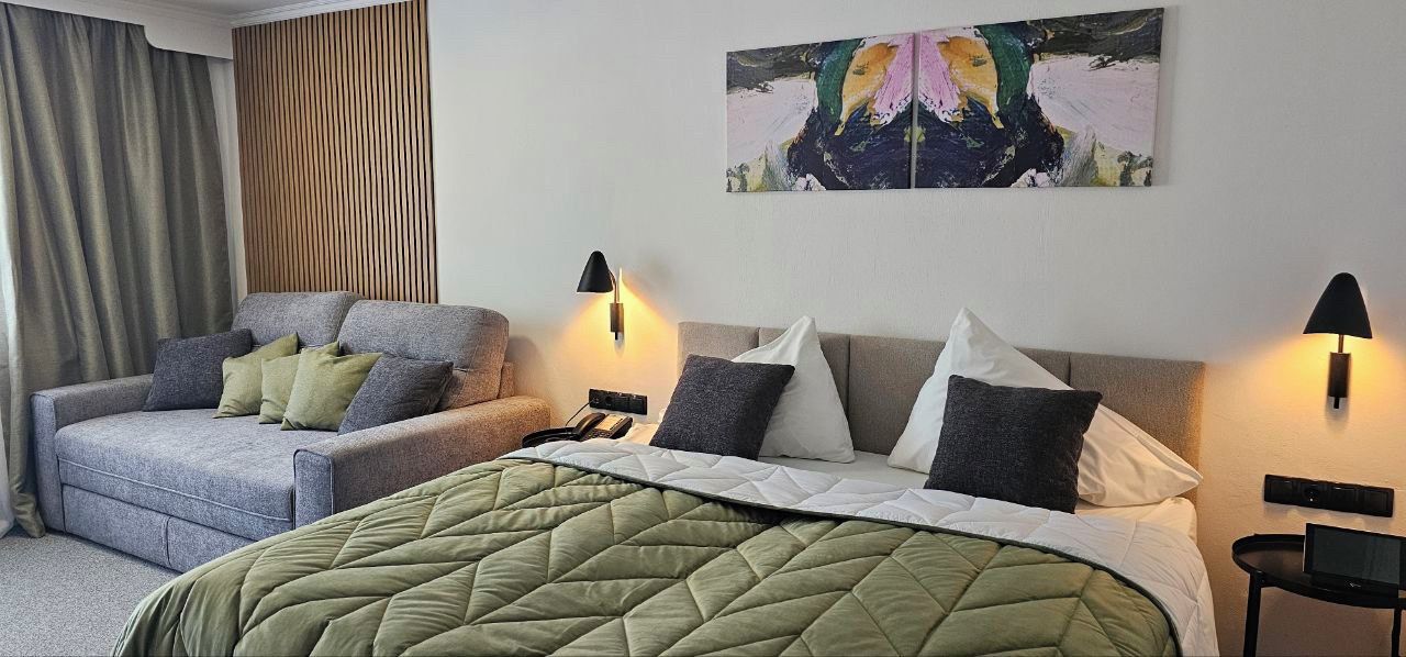 A hotel room with a bed, a sofa, two wall-mounted lamps, and a painting on the wall.