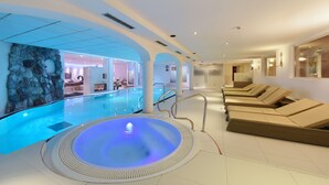A modern indoor swimming pool with a hot tub, lounge chairs, and a stone wall. - Mountains Hotel (Seefeld in Tirol)