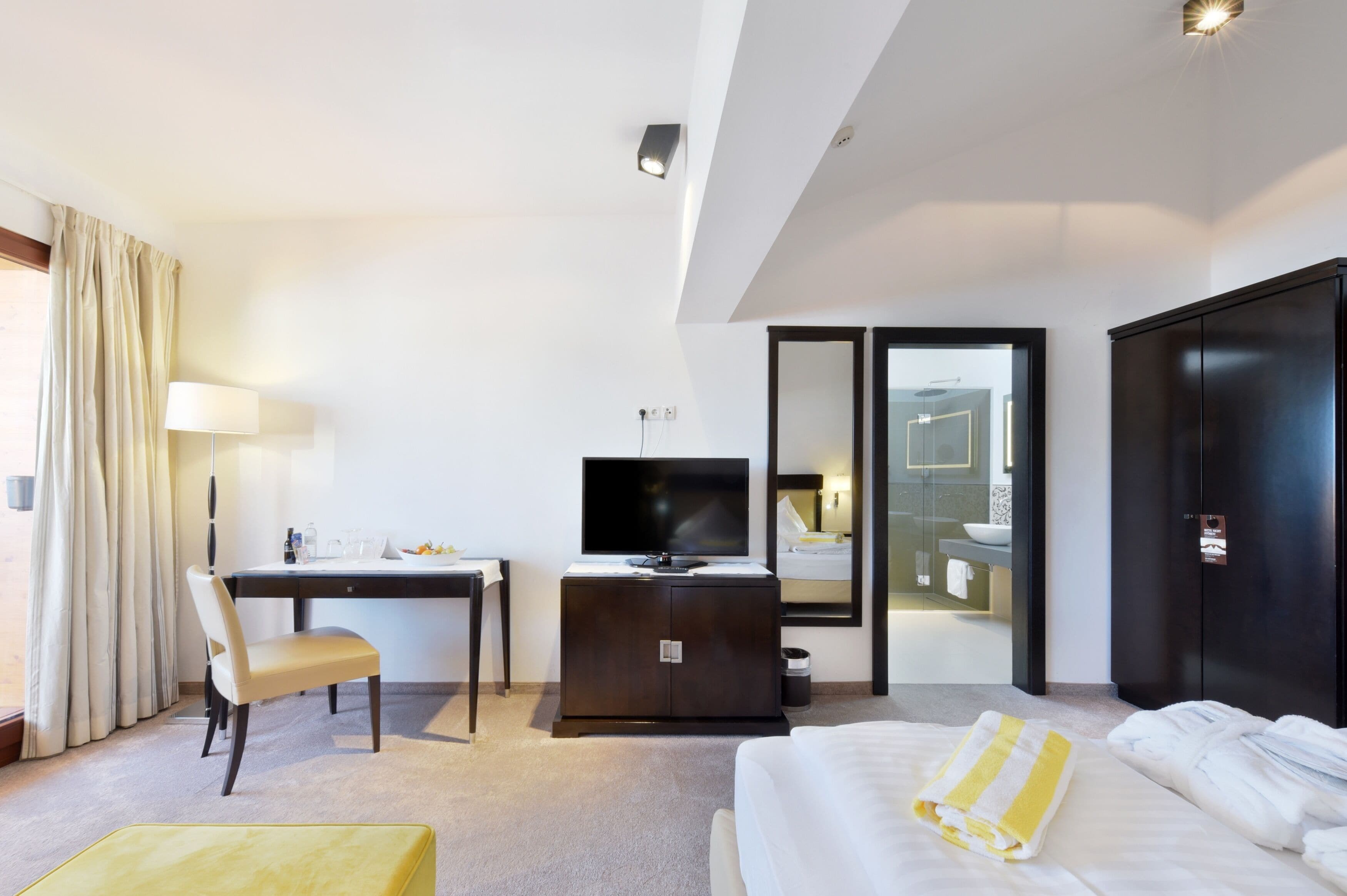 Habitación de hotel con cama, escritorio con televisor, una silla, armario y baño visible a través de una puerta de cristal.