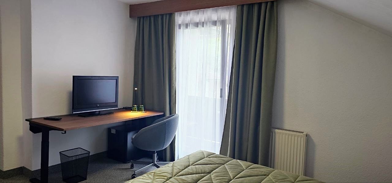 Habitación de hotel con cama, escritorio, computadora, silla y ventana con cortinas.