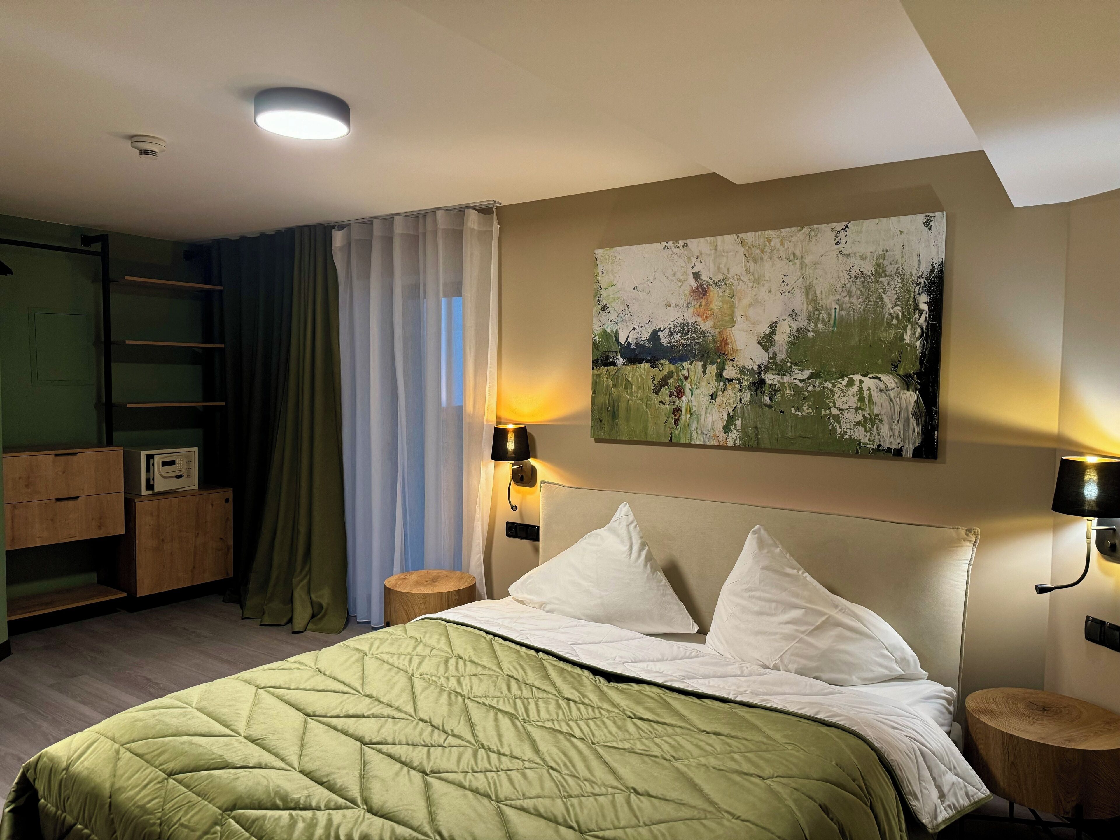 A hotel room with a bed, a painting on the wall, and a desk with a lamp.