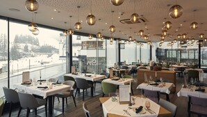 A restaurant with round pendant lights, tables set for dining, and large windows offering a view of a snowy landscape. - Mountains Hotel (Seefeld in Tirol)