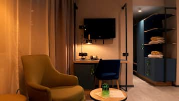 Una habitación de hotel con un sillón verde, una mesita redonda con una botella verde, un escritorio con una computadora, una silla azul y un televisor montado en la pared.