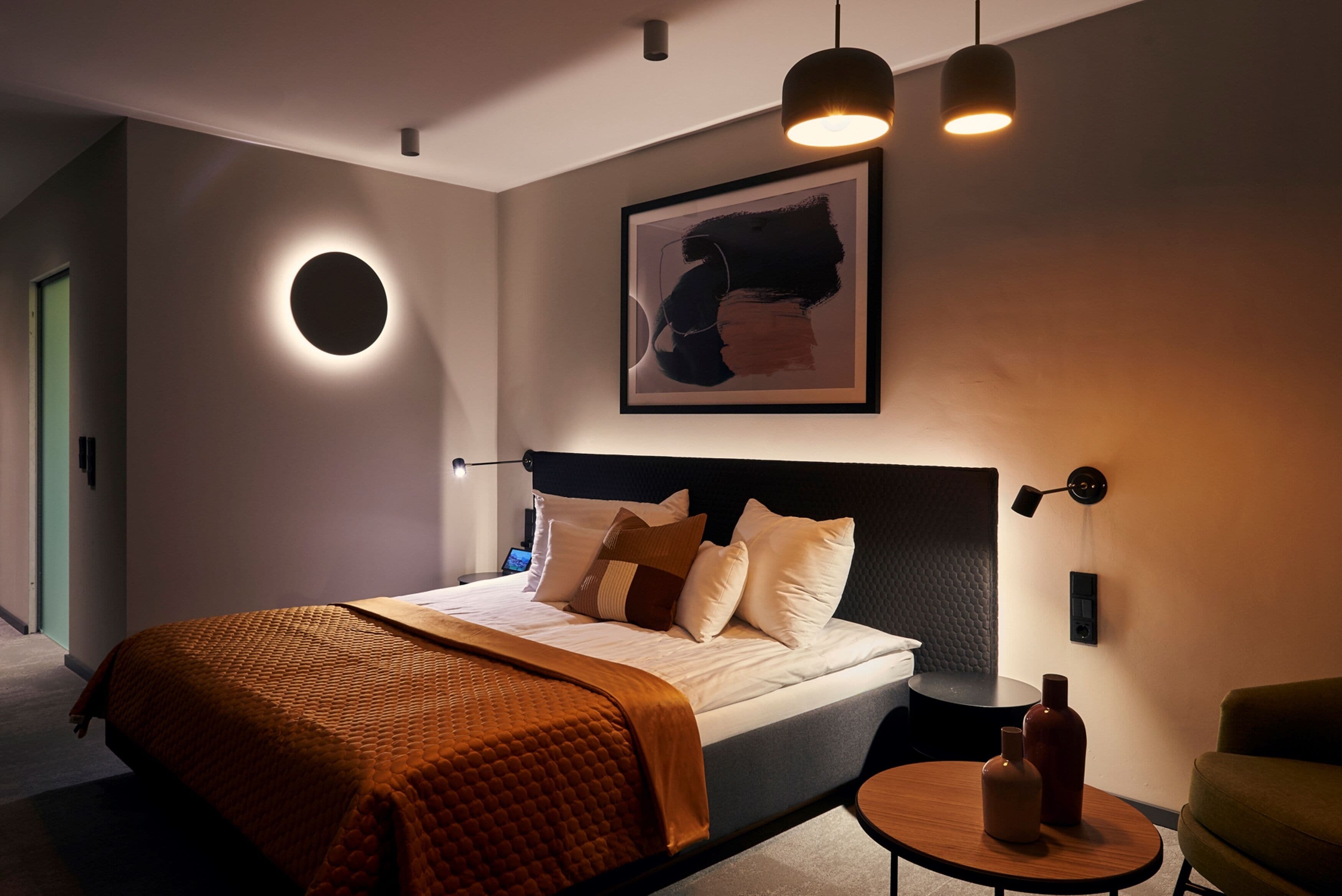 Un dormitorio moderno con una cama grande, una lámpara de pared redonda, un cuadro abstracto enmarcado y una mesita redonda con botellas.