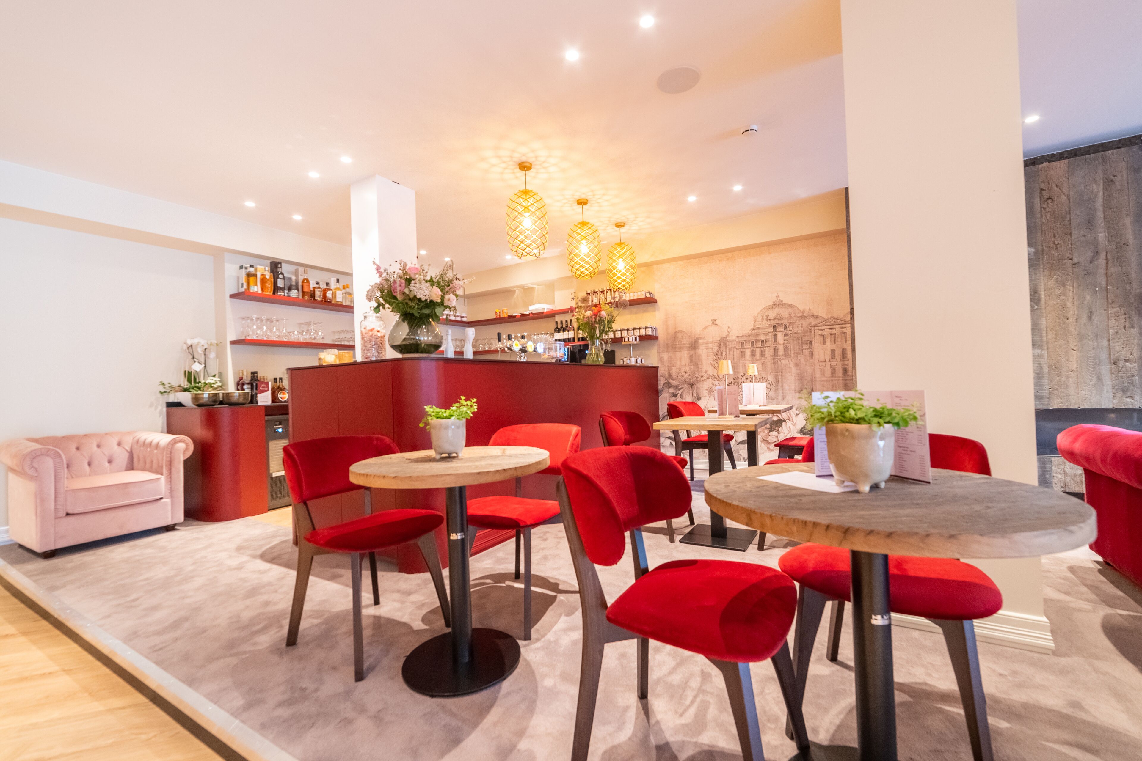 A modern bar area with red chairs and round tables, a pink sofa, and a bar counter with various bottles and glasses.
