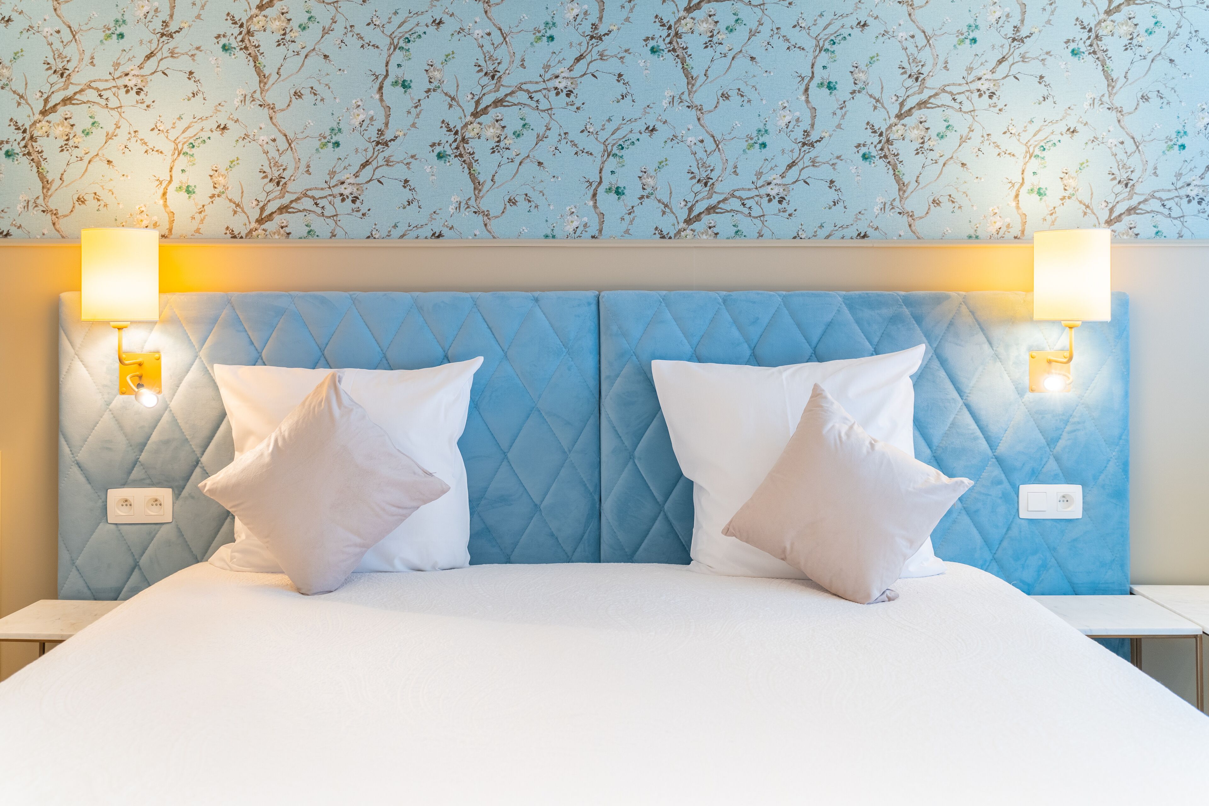 Una cama con cabecera acolchada, dos lámparas de noche y un estampado floral en el papel tapiz.