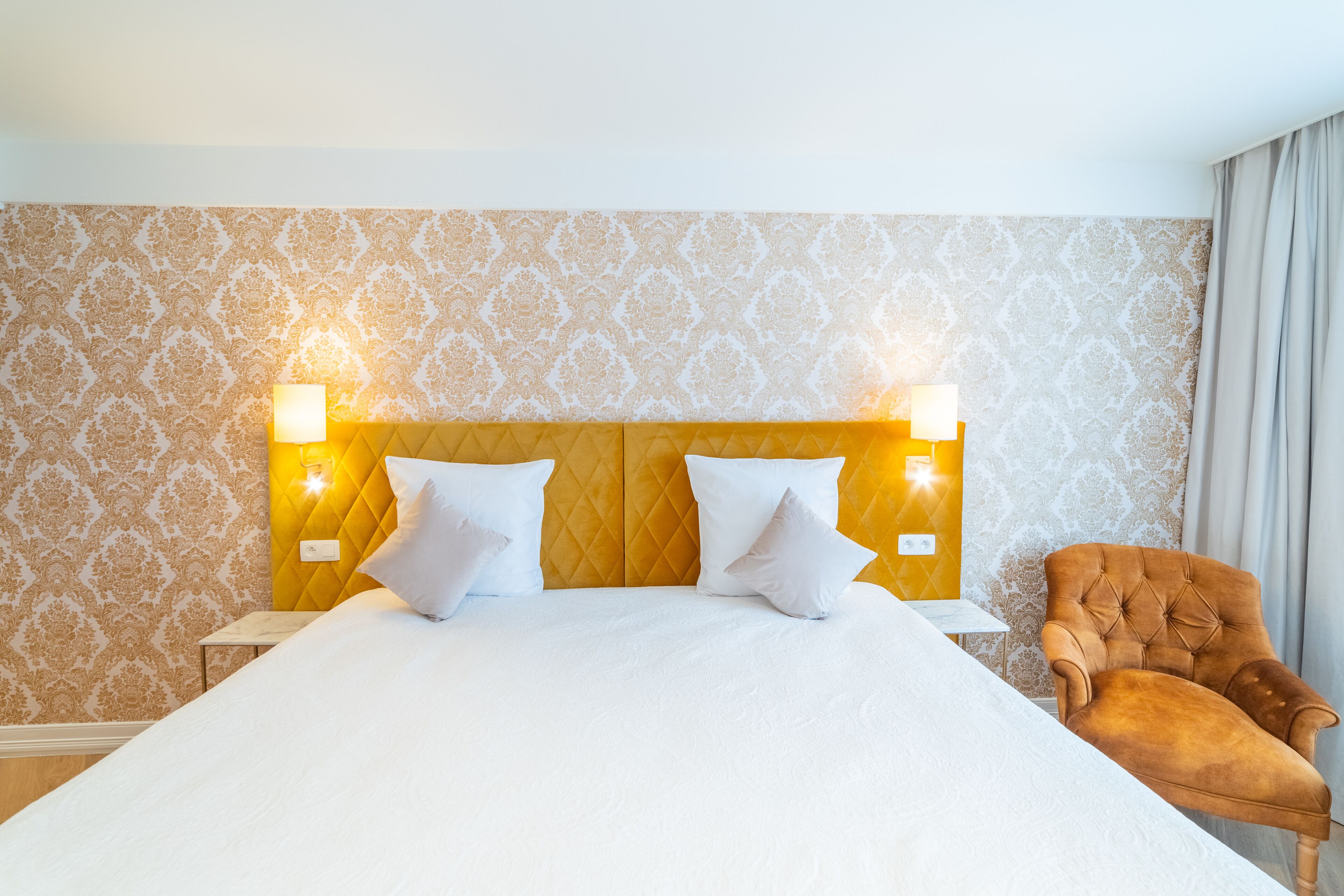 A hotel room with a large bed, two bedside lamps, a tufted headboard, and a brown armchair.