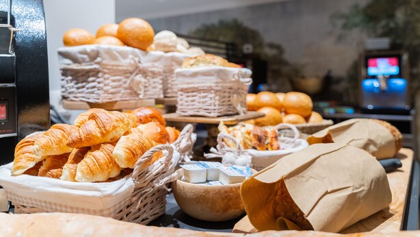 Diversas opciones de panadería y productos horneados expuestos en cestas y sobre una encimera, como croissants y panecillos.