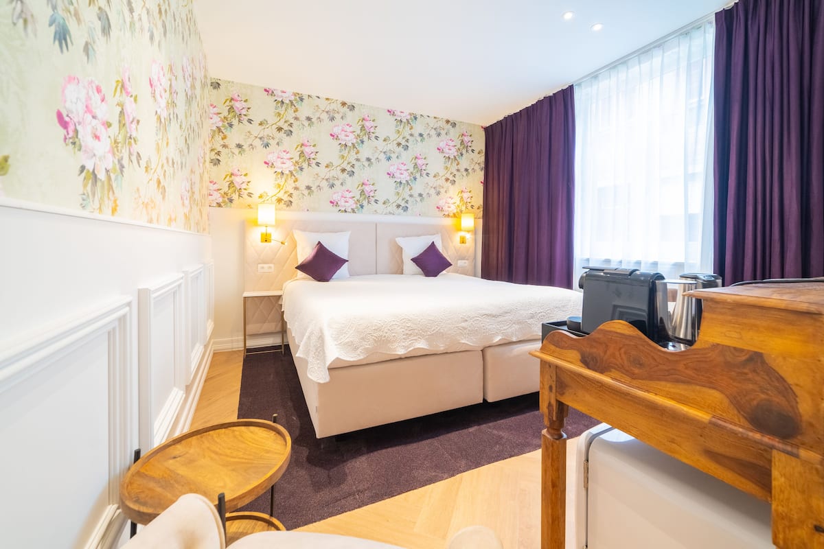 A hotel room with a large bed, a wooden desk, a small round table, and a wall with floral wallpaper.