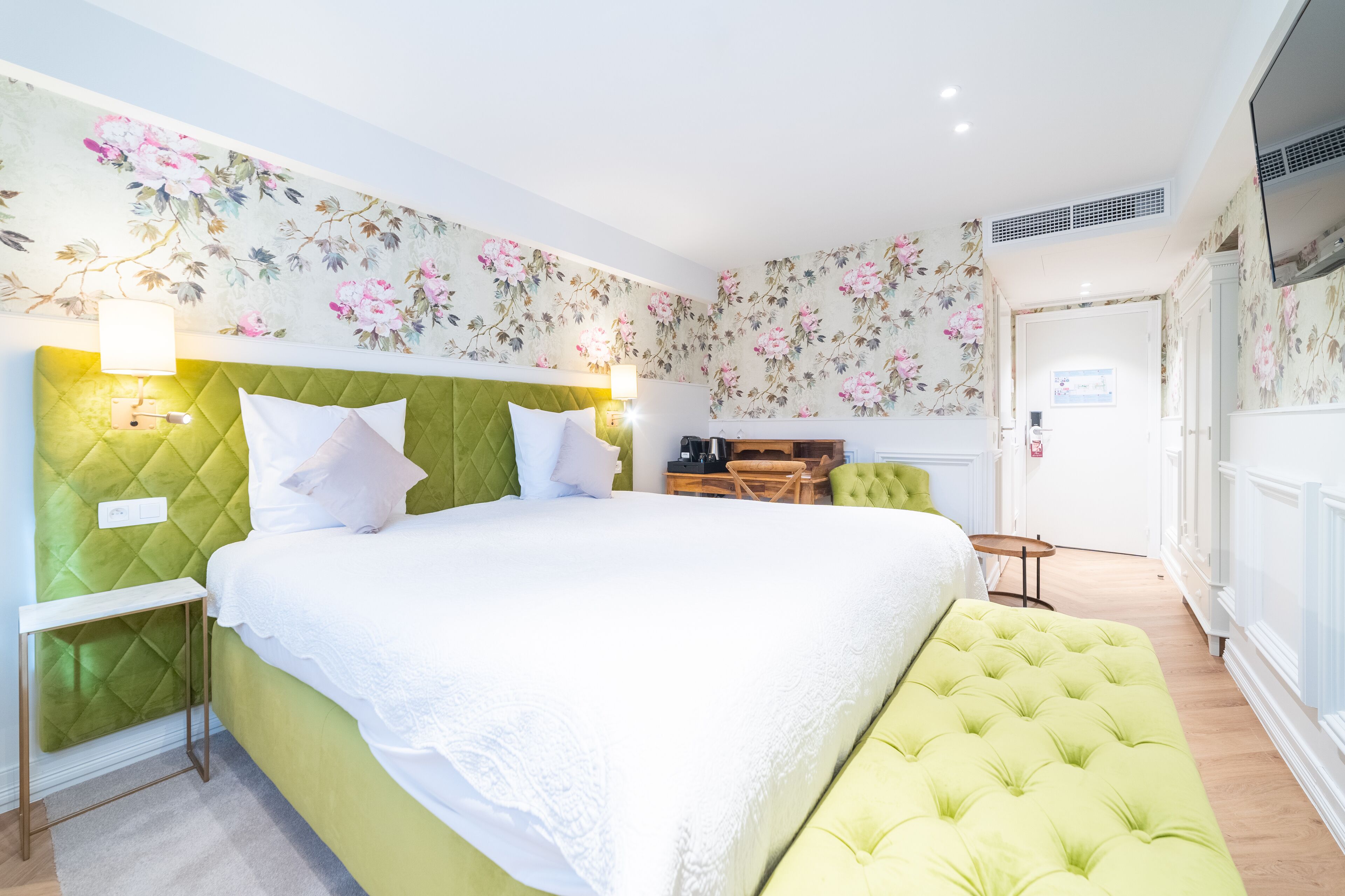 A hotel room with a green upholstered headboard, a large bed with white linens, a small table, and a green ottoman.