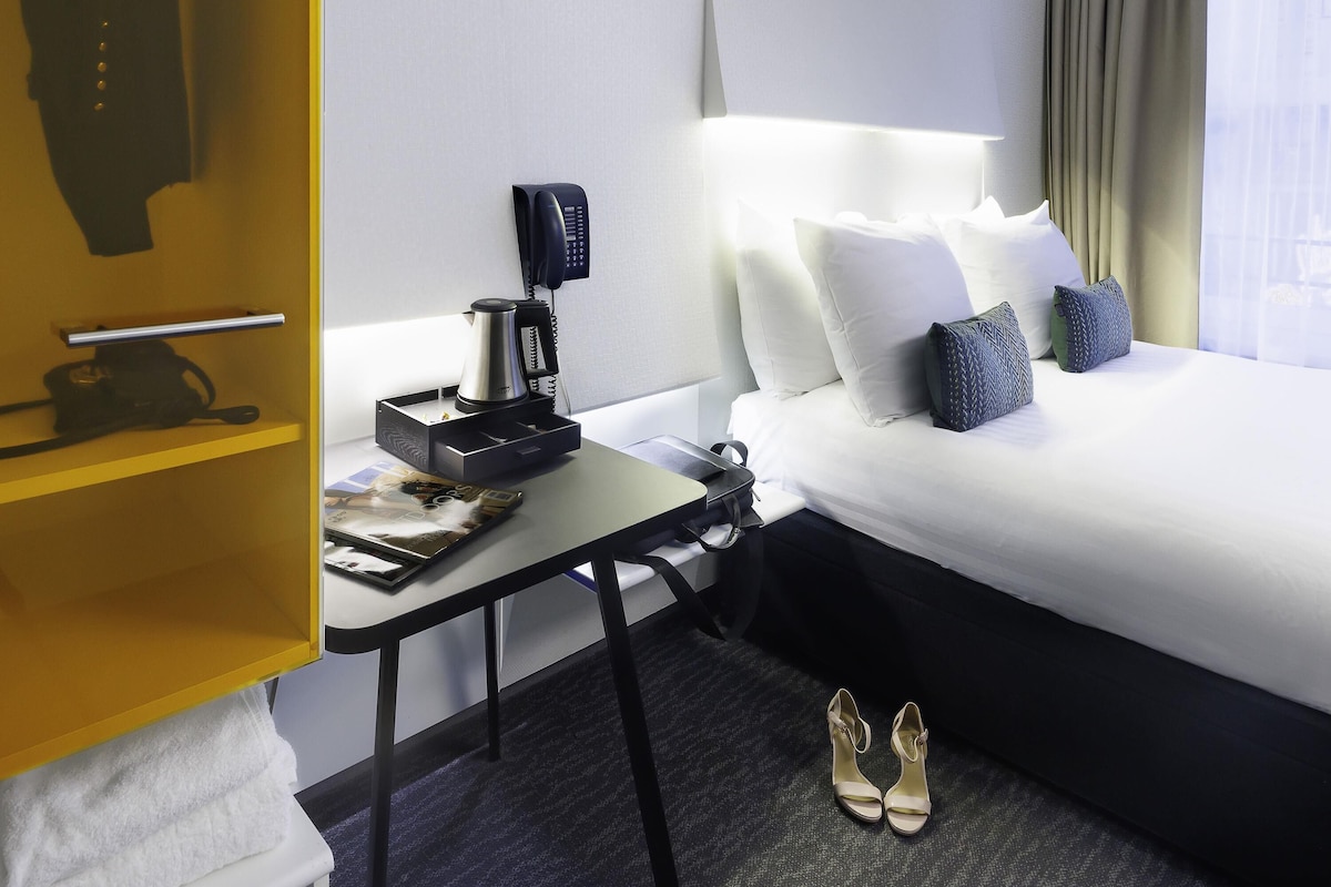 Une chambre d’hôtel avec un lit soigneusement fait, une machine à café, un téléphone et une paire de chaussures posées sur le sol.