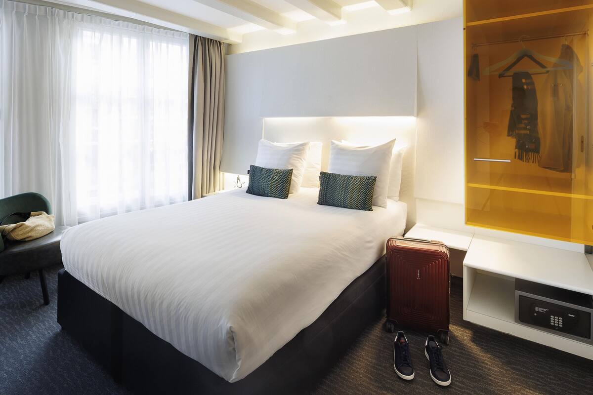 A hotel room with a large bed, a bedside table with a lamp, a wardrobe, and a suitcase on the floor.