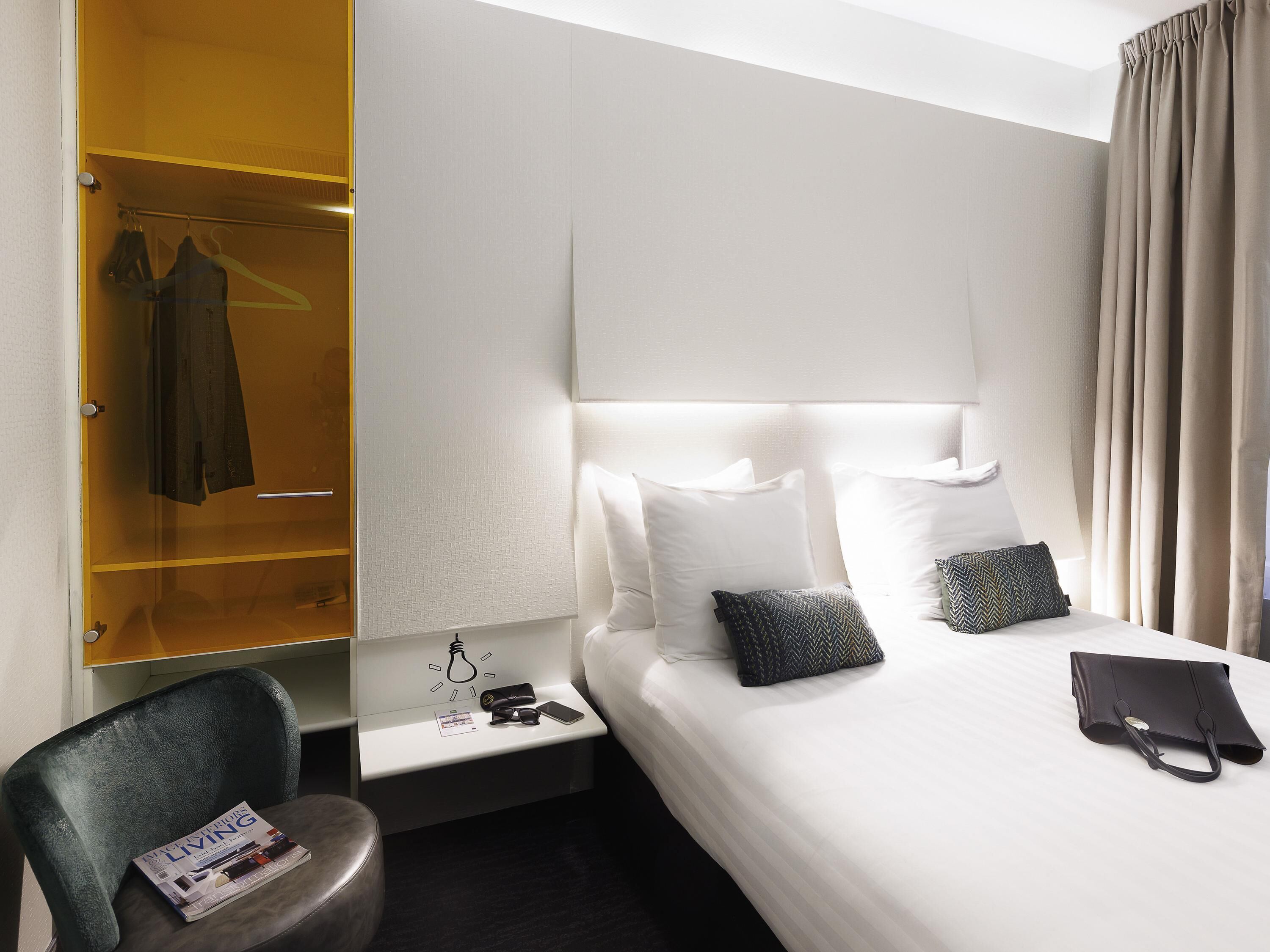 Una habitación de hotel con cama, mesita de noche con lámpara, una silla, armario y una revista sobre la silla.