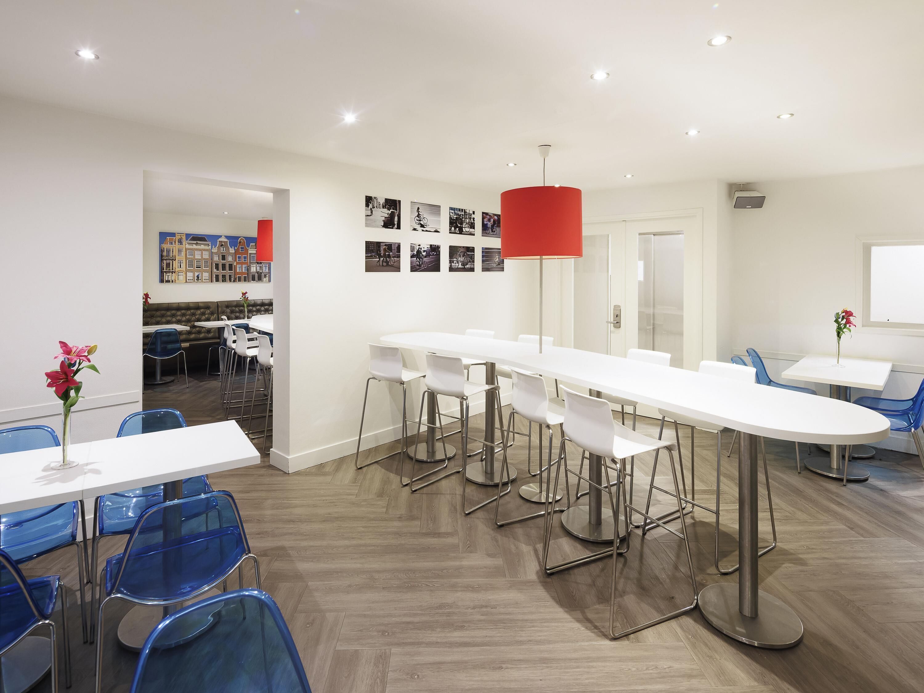 A modern dining area with white tables and blue chairs, a red pendant light, and a wall with black and white photographs.