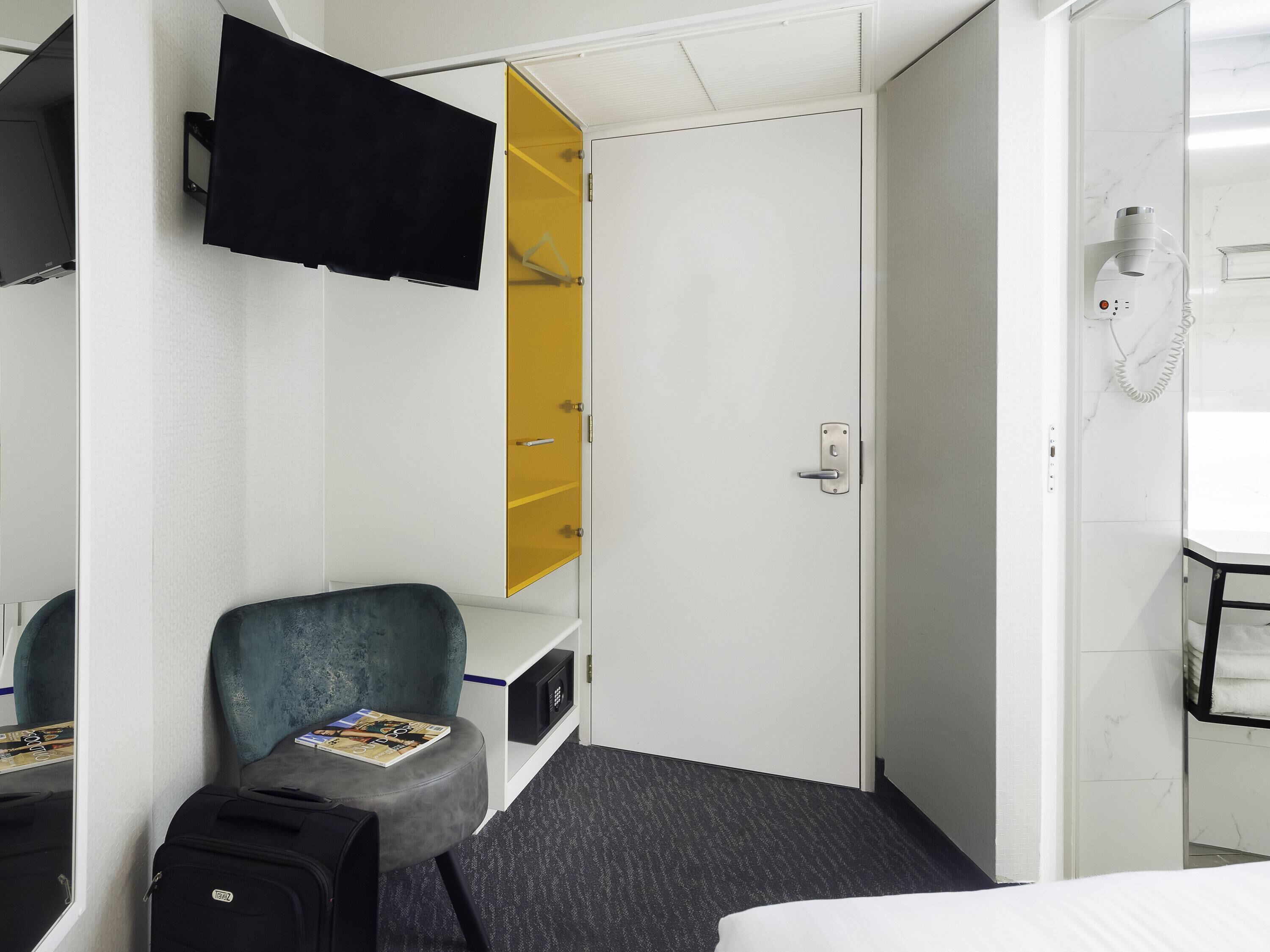 A hotel room with a bed, a desk with a computer, a chair, a suitcase, a wardrobe with a mirror, and a telephone.