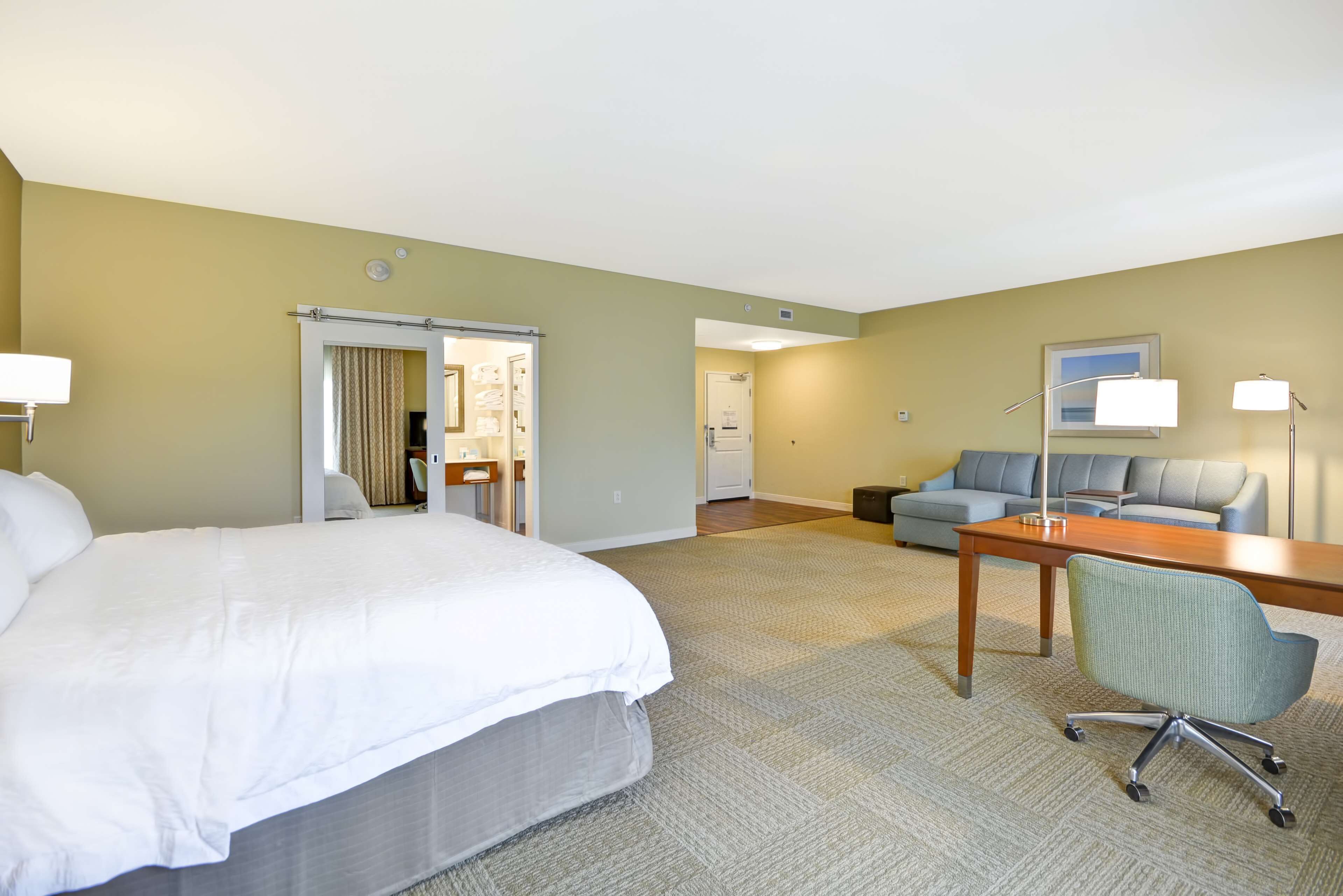 A hotel room with a large bed, a desk with a chair, a sofa, and a door leading to a bathroom.