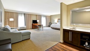 A hotel room with a sofa, a desk, a television, a bed, a bathroom with a sink, and a large mirror. - Hampton Inn & Suites Charleston Airport (North Charleston)