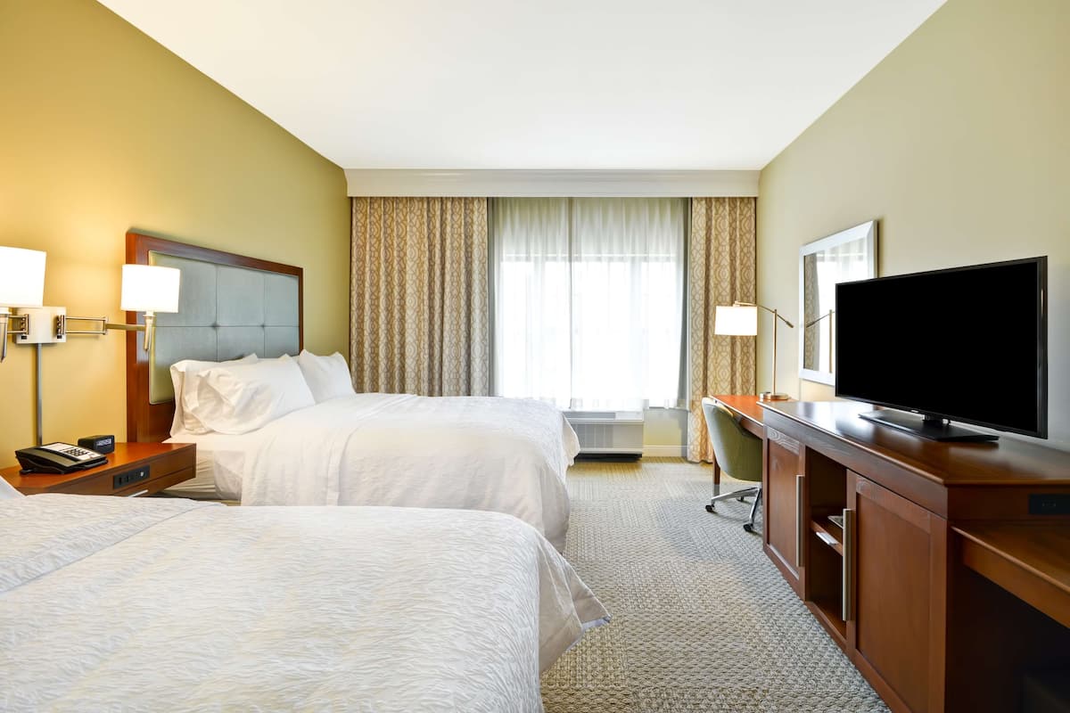 A hotel room with two beds, a desk, a television, and a window with curtains.