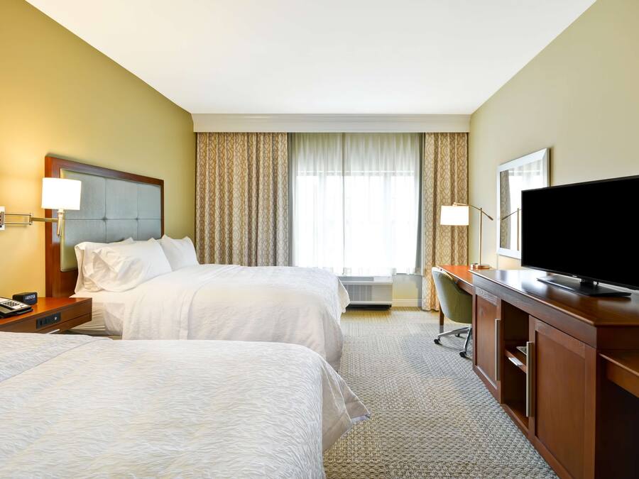 A hotel room with two beds, a desk, a television, and a window with curtains.