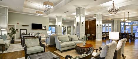 A spacious lobby with a large sofa, armchairs, and a coffee table. There are multiple chandeliers and a television mounted on the wall.