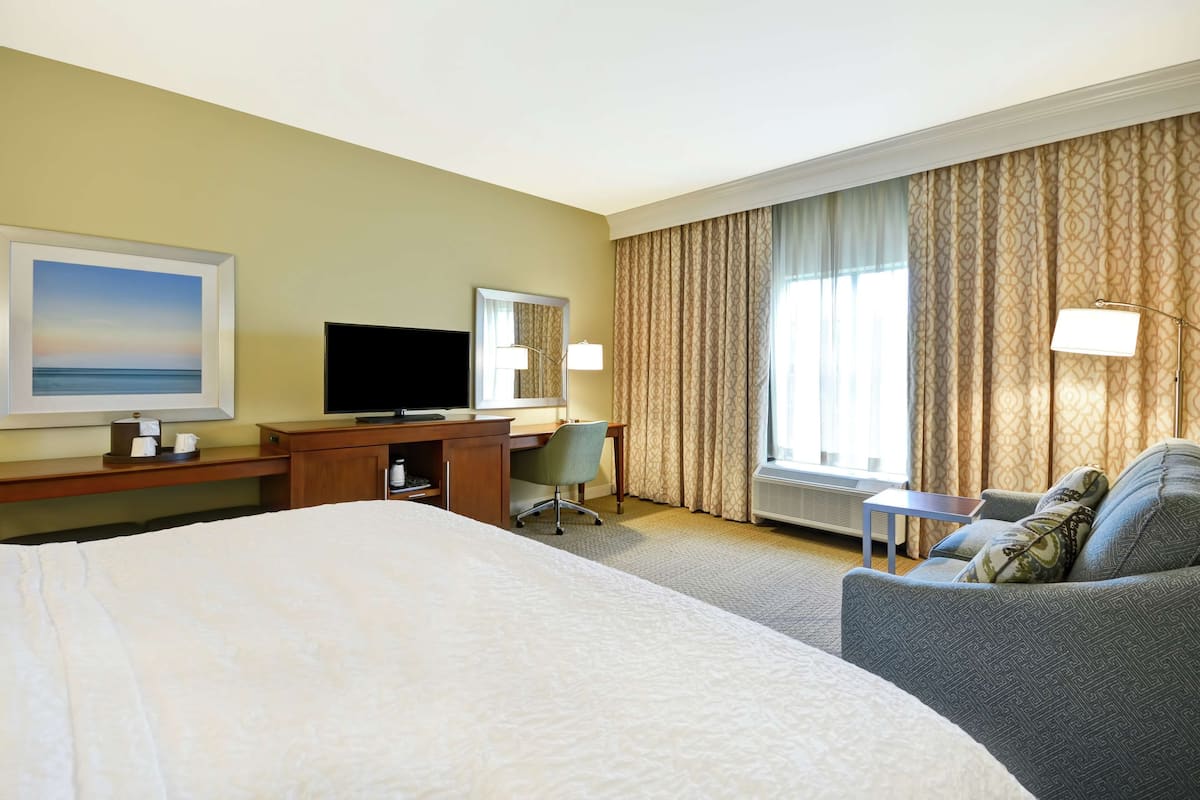 A hotel room with a large bed, a desk with a flat-screen TV, a chair, a sofa, and a window with curtains.