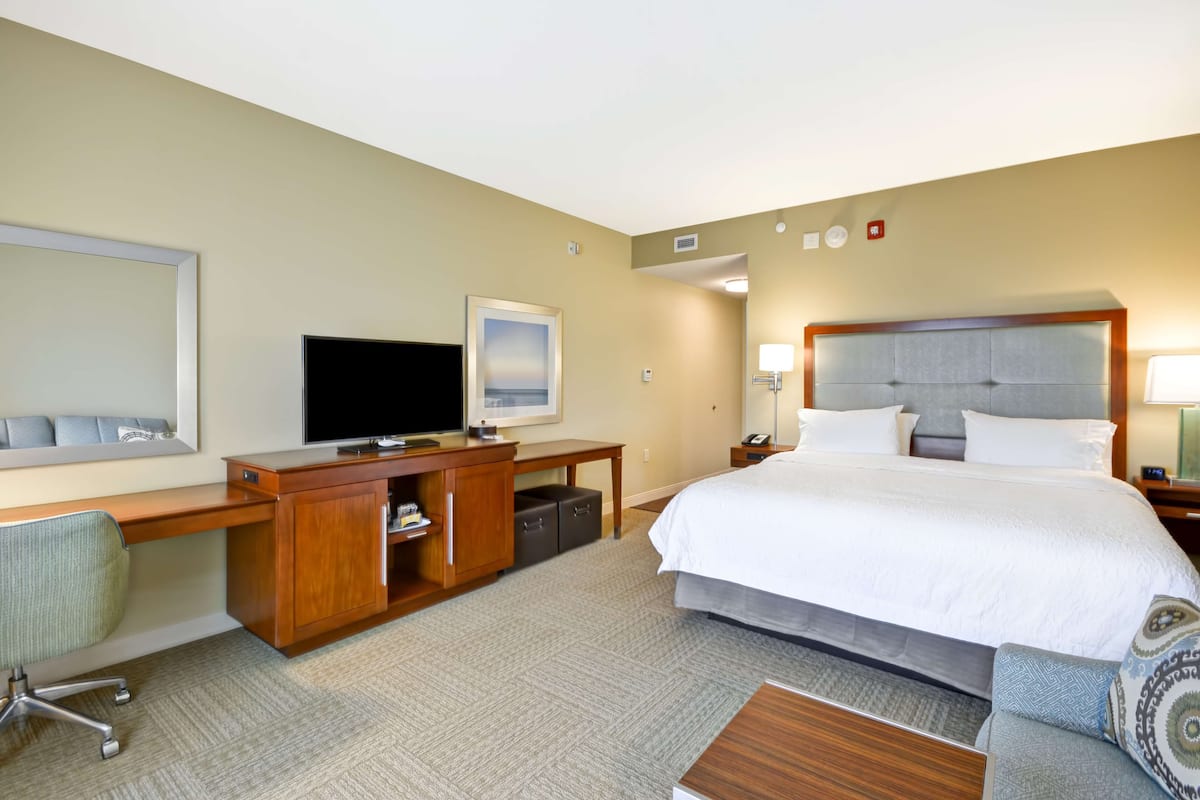 A hotel room with a large bed, a desk with a flat-screen TV, a chair, a small table, and a nightstand with a lamp.