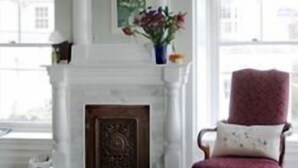 Una habitación con chimenea blanca, un sillón rojo y un jarrón con flores sobre la repisa.