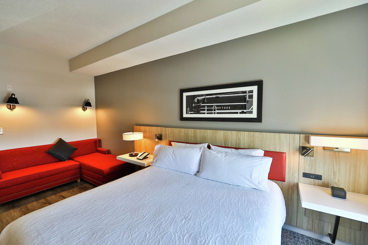 A hotel room with a large bed, a red sofa, a small table with a lamp, and a wall-mounted picture.