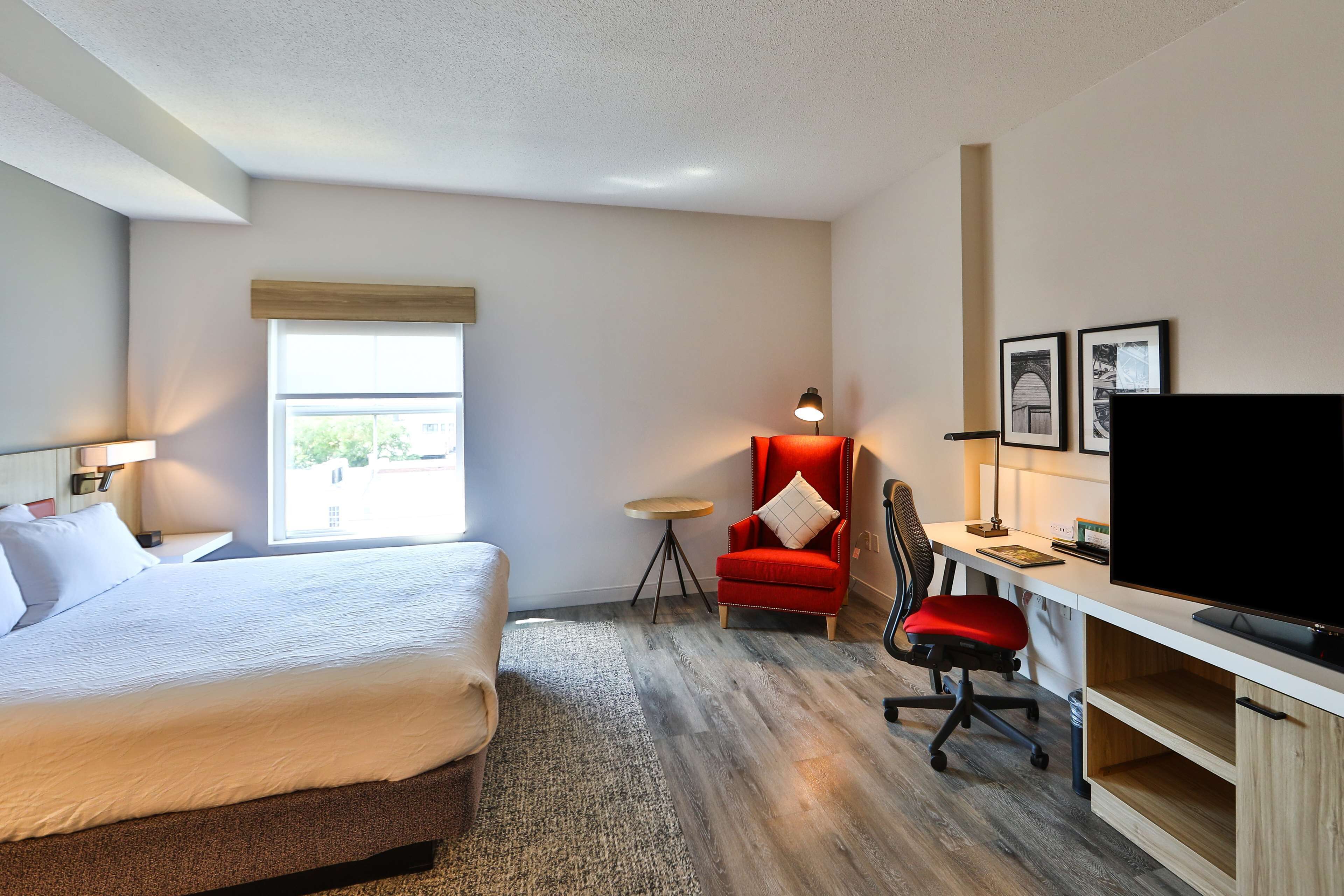 A hotel room with a bed, a desk with a TV, a red chair, and a small table.