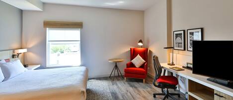 A hotel room with a bed, a desk with a TV, a red chair, and a small table.