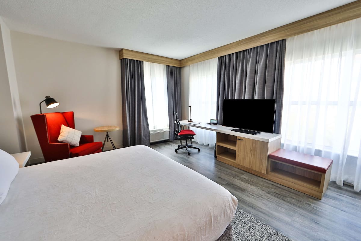 A hotel room with a large bed, a desk with a TV, a red chair, and a small table.