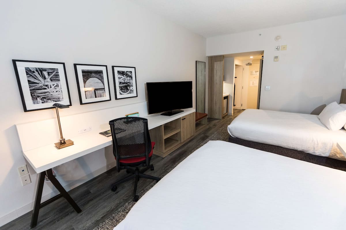 A hotel room with a white bed, a desk with a computer, a TV, and framed pictures on the wall.