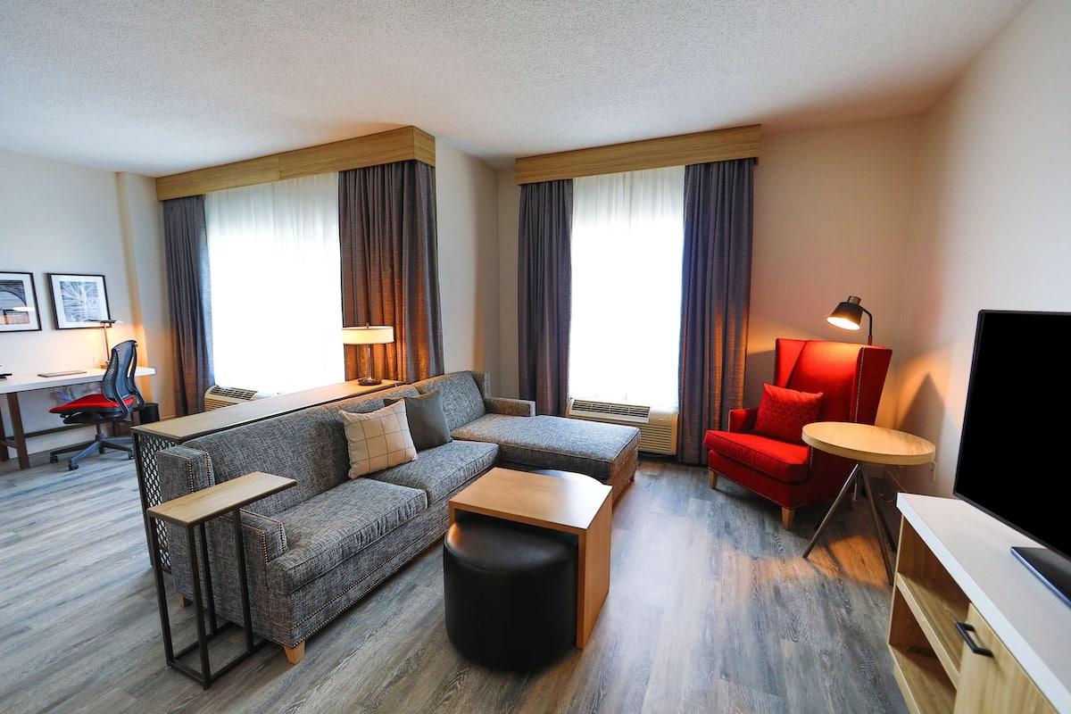 A modern hotel room with a grey sofa, a wooden coffee table, a red armchair, and a TV.