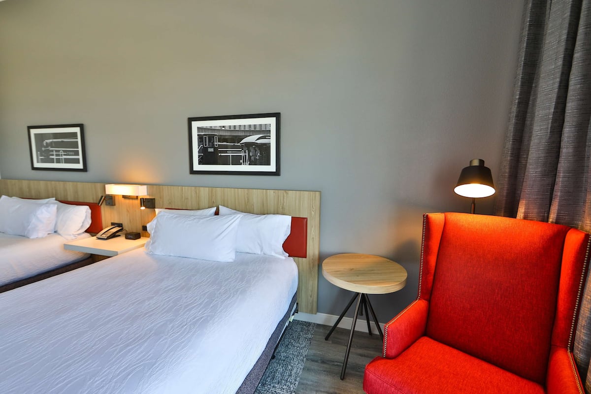 A hotel room with two beds, a red armchair, a small round table, and two framed pictures on the wall.