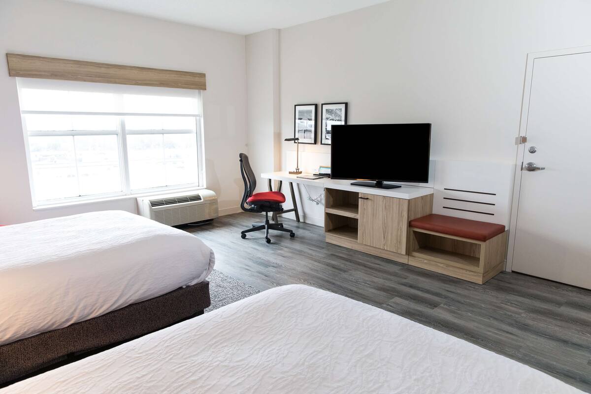 A hotel room with two beds, a desk with a TV, a chair, and a bench.
