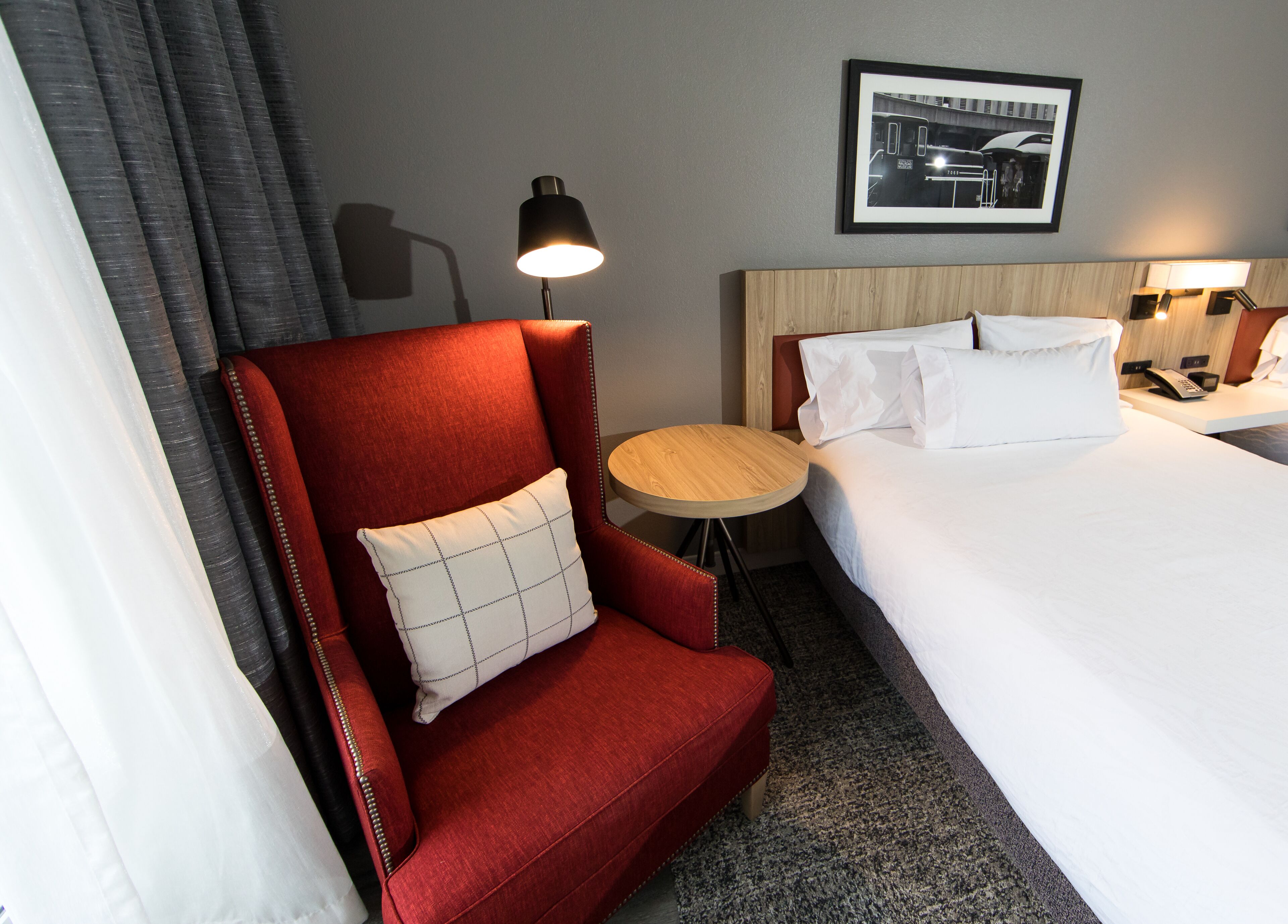 Une chambre d’hôtel avec un fauteuil rouge, une petite table ronde, un lit avec du linge de maison blanc et un tableau encadré au mur.