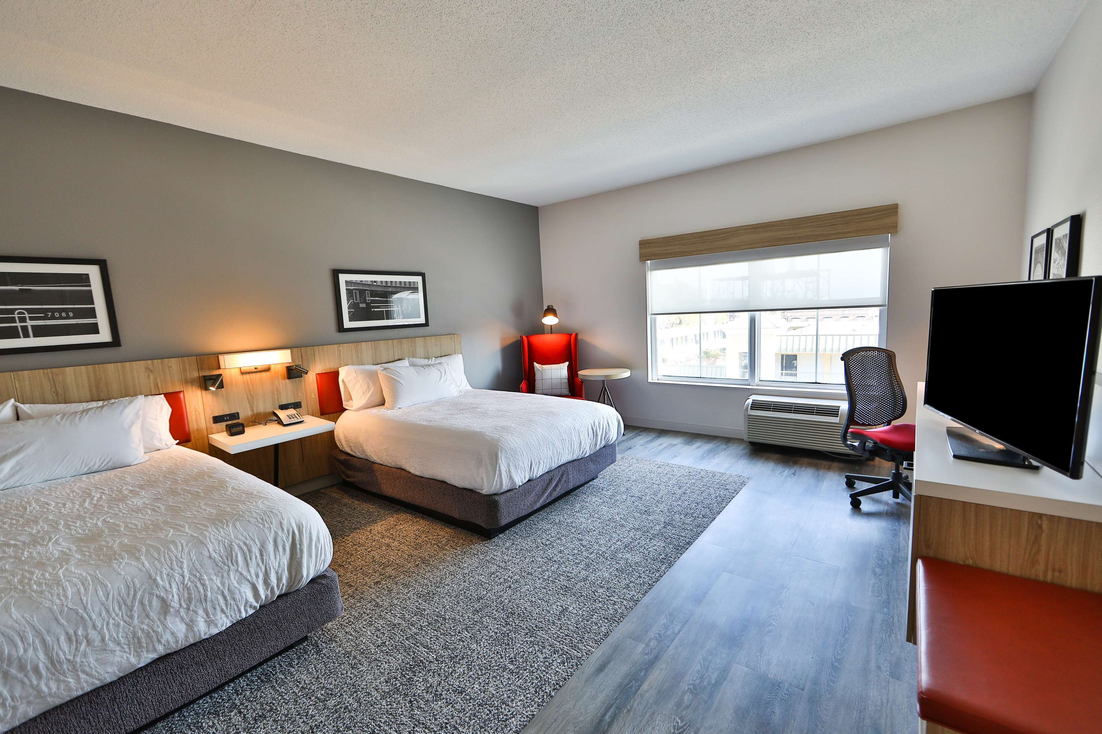 A hotel room with two beds, a desk with a flat-screen TV, a chair, a lamp, and framed pictures on the wall.