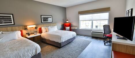 A hotel room with two beds, a desk with a flat-screen TV, a chair, a lamp, and framed pictures on the wall.