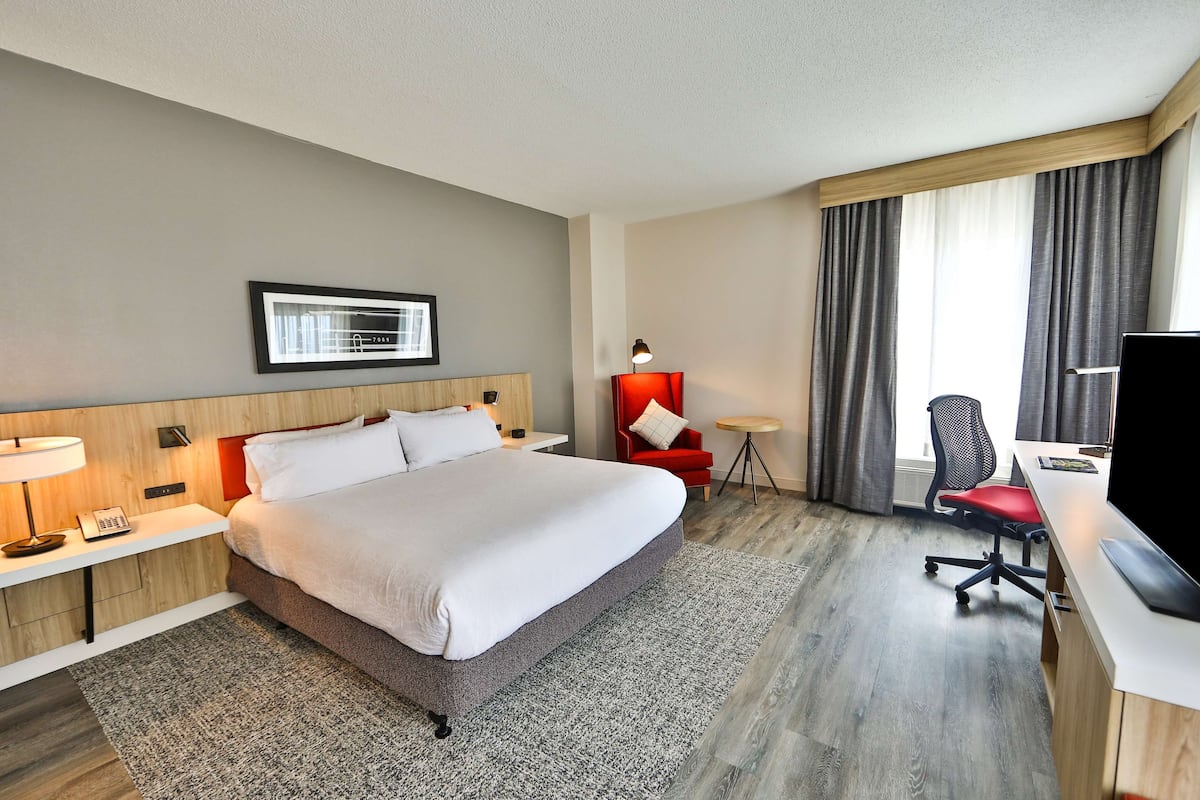 A hotel room with a large bed, a desk with a computer, a chair, a red armchair, and a window with curtains.