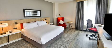 Une chambre d’hôtel avec un grand lit, un bureau avec un ordinateur, une chaise, un fauteuil rouge et une fenêtre avec des rideaux.
