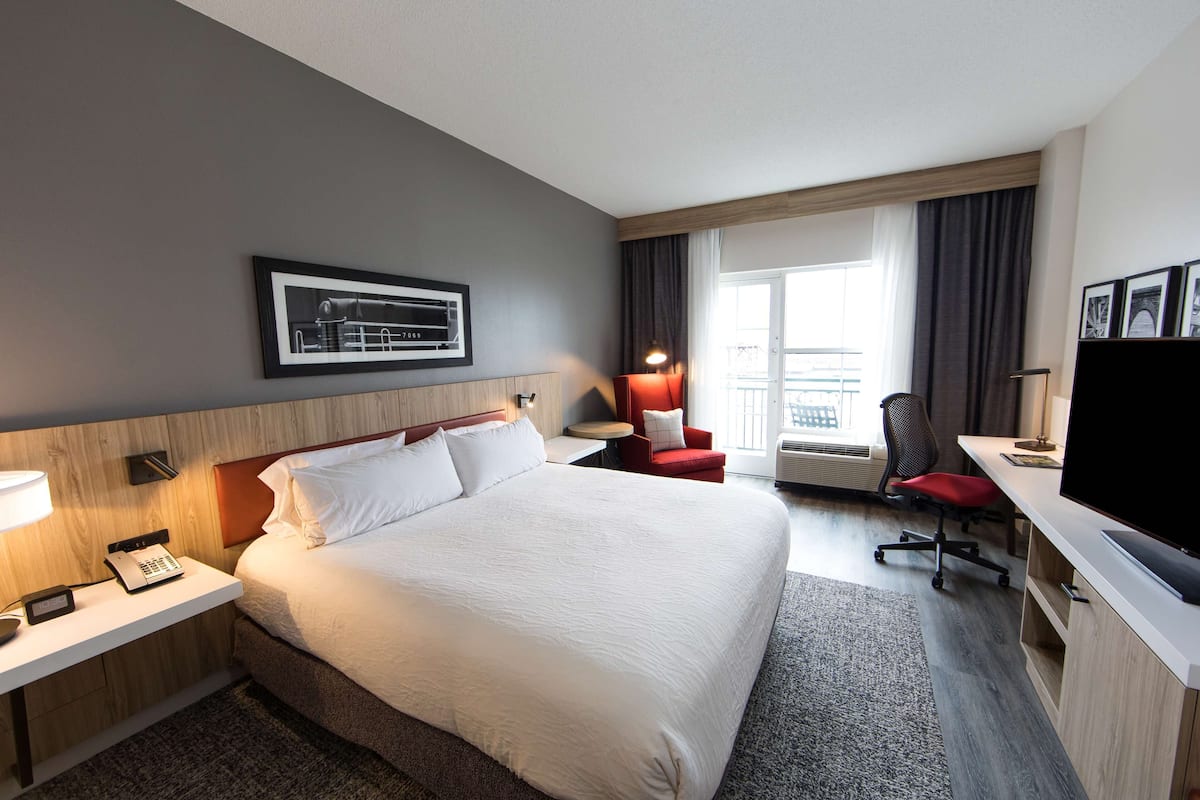 A hotel room with a large bed, a desk with a computer, a chair, a TV, and a window with curtains.