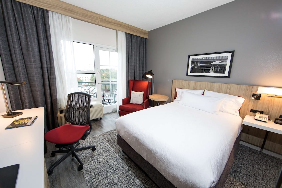 A hotel room with a large bed, a desk with a chair, a small round table, and a balcony with a railing.