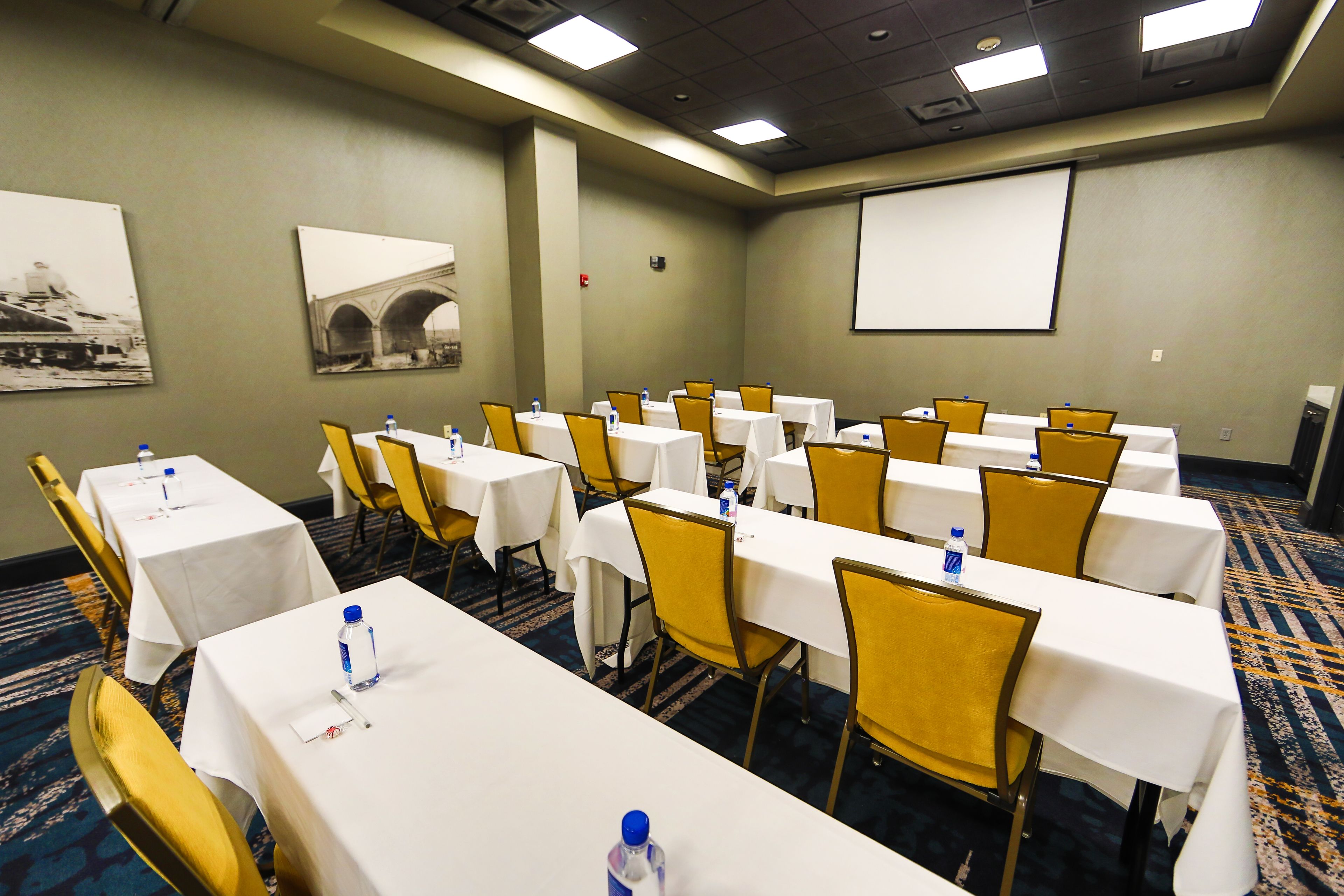 Una sala de conferencias con varias mesas y sillas, equipada con botellas de agua y una pantalla de proyector en la pared.