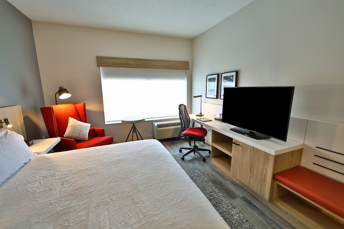 A hotel room with a bed, a desk with a TV, a red chair, a lamp, and a window.