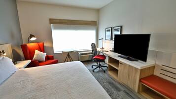 A hotel room with a bed, a desk with a TV, a red chair, a lamp, and a window.