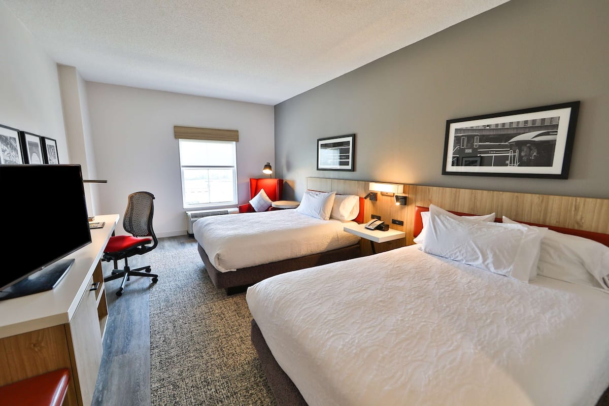 A hotel room with two beds, a desk with a TV, a chair, and a window with a view of the city.