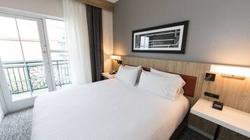 Habitación de hotel con una cama grande, una mesita de noche con un reloj, un cuadro en la pared y una ventana con cortinas.