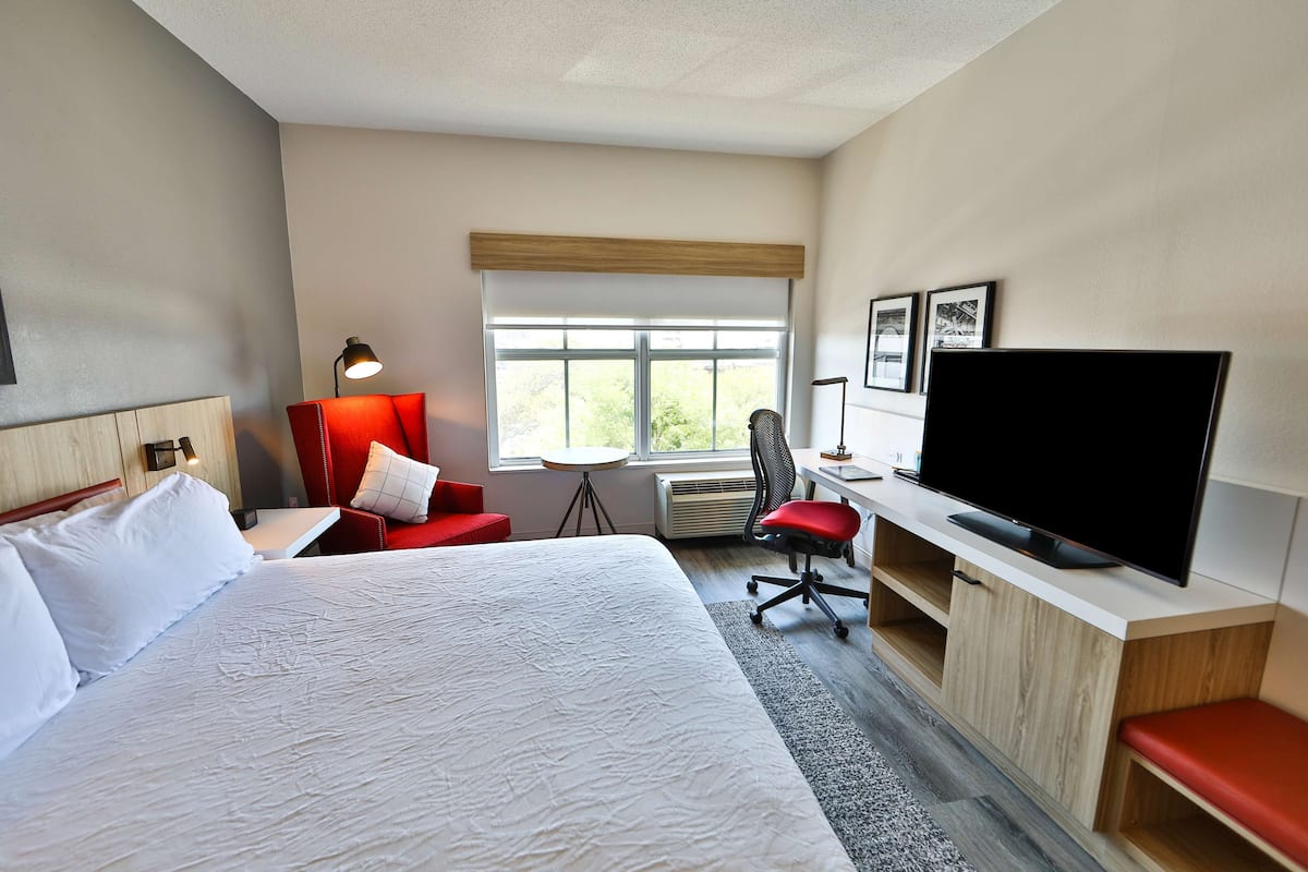 A hotel room with a large bed, a desk with a flat-screen TV, a red chair, and a window with a view of greenery.